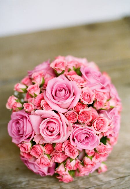 A round bouquet arrangement featuring large, soft pink roses prominently in the center, surrounded by smaller pink rosebuds and blooms, all with lush green foliage. The flowers are fresh, with velvety