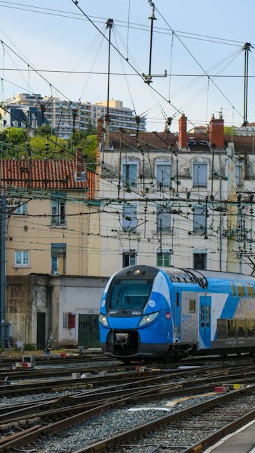 The image shows a modern blue and white train arriving at or departing from a railway station with visible overhead electric lines. In the background, there are multi-storey residential buildings with