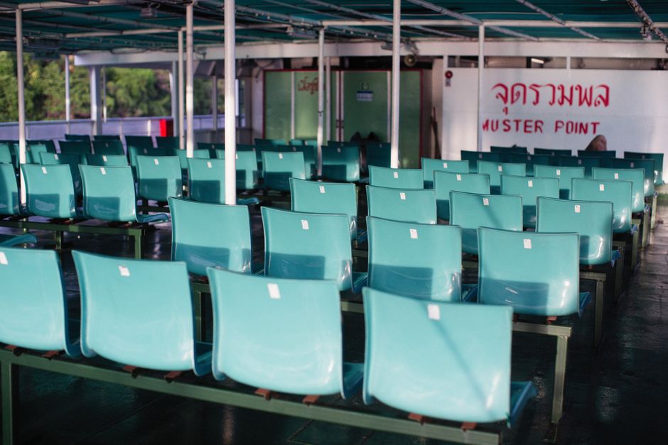 Empty turquoise plastic seats arranged in rows on a covered outdoor waiting area, with a white wall and red Thai script and English text reading 'MUSTER POINT' in the background. The seating is illumi