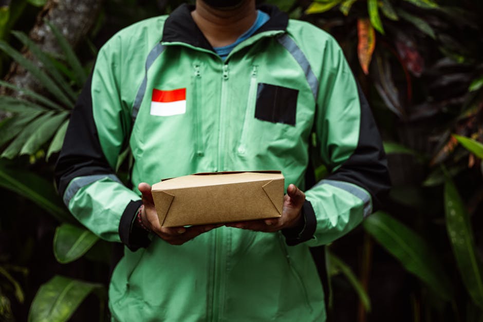 A delivery person dressed in a high-visibility green jacket with reflective grey stripes, holding a rectangular parcel wrapped in plain brown paper with neatly folded corners, standing outdoors amidst