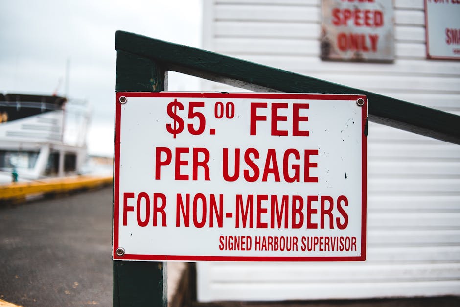 A white rectangular sign with bold red lettering displaying a '$5.00 FEE PER USAGE FOR NON-MEMBERS' message, attached to a dark green wooden post with visible screws. The background shows part of a ma