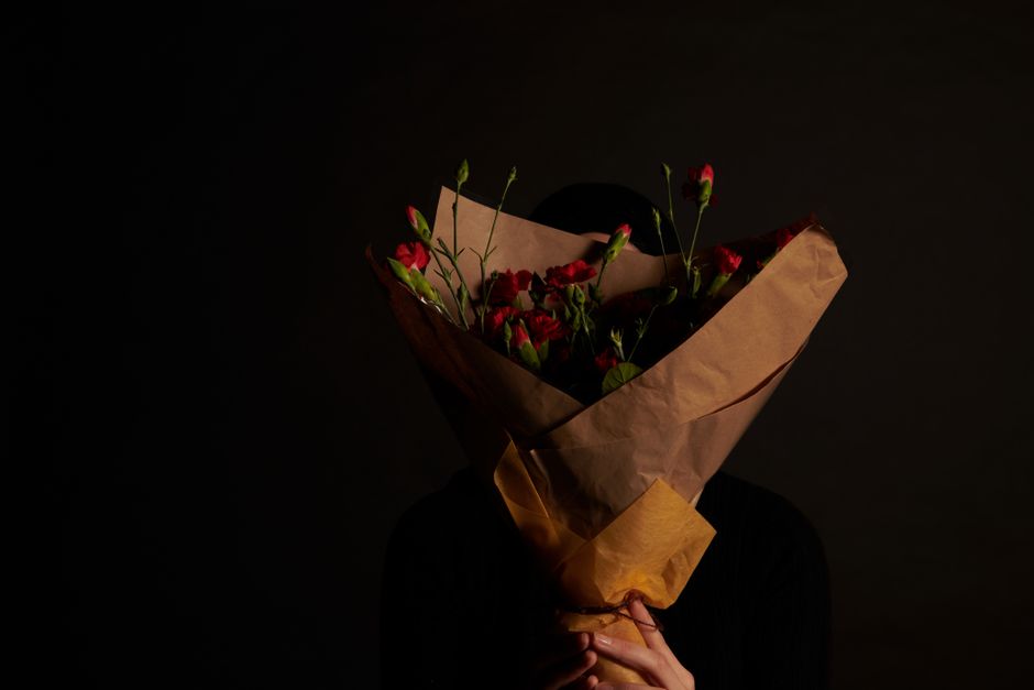 A person holding a bouquet of fresh red and pink flowers, including carnations, wrapped in light brown kraft paper with a beige paper band at the base, against a dark background. The bouquet is held c