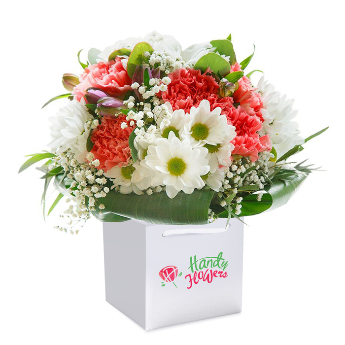 Set against a bright white background, this exquisite Collier Row floral arrangement is showcased within a neat, square white gift bag-its sides crisp and its ribbon handles strong, ideal for convenient gifting. The bouquet's striking palette unites coral-pink and deep red carnations, renowned for their soft, ruffled petals, with a scattering of daisy-like white chrysanthemums whose lime-green centers convey a touch of early spring vitality. Dainty clouds of white baby's breath are sprinkled throughout, creating a delicate, cloud-like texture that lightens the whole effect. The blooms are closely encircled by lush, deep green foliage-broad, glossy leaves form an organic collar to the bouquet, much like the woodland edges around Collier Row's parks. Peeking from the arrangement are several purple-tinted, closed buds, adding mystery and layered depth-subtle reminders of newly budding flowers along White Hart Lane. The entire composition is arranged with a sense of fullness and natural abundance, carefully balanced and inviting. Each stem appears fresh and thoughtfully positioned, a testament to the artistry and precision of Collier Row's local florist community. Perfect for delivering warm wishes-from milestone birthdays to spontaneous celebrations-this bouquet captures both seasonal colour and sincere sentiment.