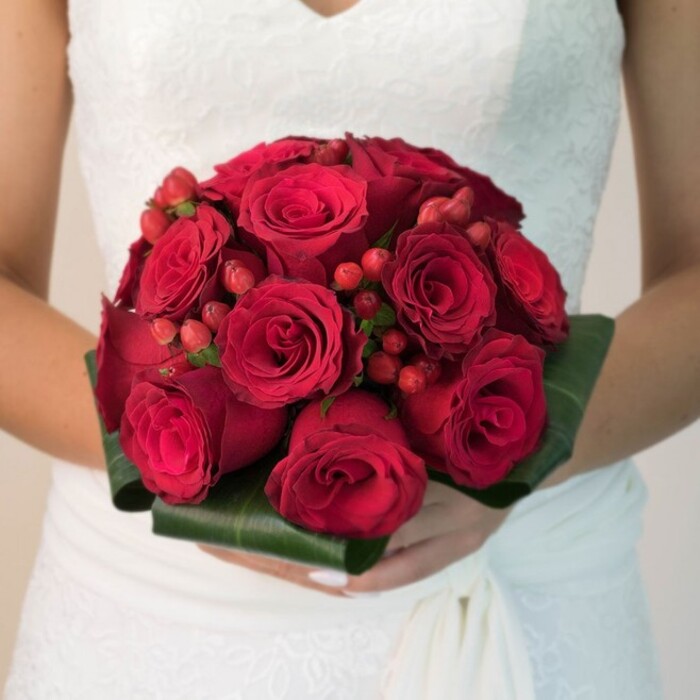 This striking image showcases a bride, captured from the waist up, reverently holding an opulent bouquet in her hands-a scene easily imagined at a wedding near Lawns Park or Central Parade in Collier Row. Her crisp white dress features fine lacework along the bodice and transitions seamlessly into a flowing skirt, the soft folds of fabric a delicate canvas for the bouquet. The floral arrangement radiates intensity, composed of dozens of velvety, deep ruby red roses-each with petals shaped in immaculate, intricate spirals. Interspersed among the blooms are vivid red hypericum berries, clustered in small groups, bringing both texture and a spirited brightness to the arrangement. The bouquet is tailored with a generous collar of broad, dark green leaves, their glossy sheen and perfect folds providing a natural yet sophisticated frame. Illuminated by even, diffused light, every detail-from the shimmer on a berry to the velvety surface of a rose-glows gently. The bride's neatly manicured hands underscore the bouquet's significance, and a sense of ceremony is heightened by the luxurious mood. This composition tells a tale of local Collier Row dedication and creative spirit, symbolising enduring love and the cherished traditions of wedding celebrations throughout the community.