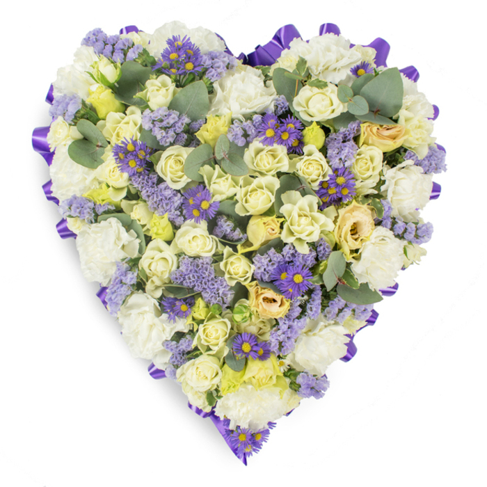 A carefully arranged heart-shaped tribute photographed on a crisp white surface, the display reads as a composed and serene memorial piece. The heart is crisply outlined with a continuous band of deep purple satin ribbon, the folded edging giving the design a defined, classical silhouette. Within, the palette is soft and restrained: abundant creamy roses in pale yellow and ivory, ranging from tight buds to softly unfurling blossoms, create a layered, tactile centre that conveys both delicacy and steadiness. Placed among these are clusters of white carnations whose ruffled forms add a plush, comforting fullness. Light lavender statice disperses throughout like whispers of colour, while small purple daisy-like flowers with striking yellow centres provide concentrated touches of deeper hue that prevent the arrangement from feeling one-dimensional. The scattered blue-green eucalyptus leaves offer a cool, rounded contrast to the petal shapes and hint at a subtle herbaceous scent, visually breathing between dense florals. The overall composition balances texture, colour, and form to evoke peace, respect, and lasting affection-qualities appropriate for funeral services, memorials, and graveside offerings. Seen here under soft, diffuse lighting that enhances petal translucence and the satin's sheen, this heart-shaped design would be familiar to neighbours around Collier Row Recreation Ground and those arranging tributes for services in Romford and the surrounding Havering parishes.