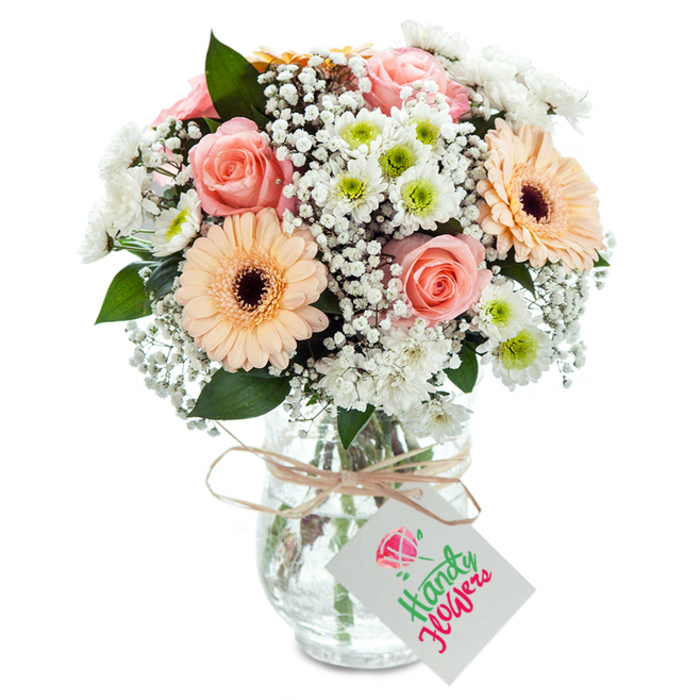 Bright sophistication finds form in this stunning bouquet, crafted in the heart of Collier Row and presented in a clear, gently textured glass vase. Romantic peach-pink garden roses are the mainstay, their unfurling petals inviting touch and conveying the gentle embrace of a close friend. Alongside, two bold peach gerbera daisies shine-robust, symmetrical petals cupping their rich brown cores. Interwoven among the larger flowers are plentiful sprays of tiny, cloudlike gypsophila and compact, daisy-like white chrysanthemums, their centers pulsing with lime green vibrance. Sparse eucalyptus or similar deep greenery peeks between flowers, offering a hint of woodland serenity much like the tree-lined paths that meander through Collier Row. The stems, visible in the clear vase, reveal attentive preparation and promise freshness. Tied simply with rustic twine, a blank white tag hangs, gracefully understated, ready to be transformed by a bespoke message. This arrangement, aglow in soft daylight, would be treasured for any occasion-its beauty a testament to the artistry found at Collier Row Flowers.