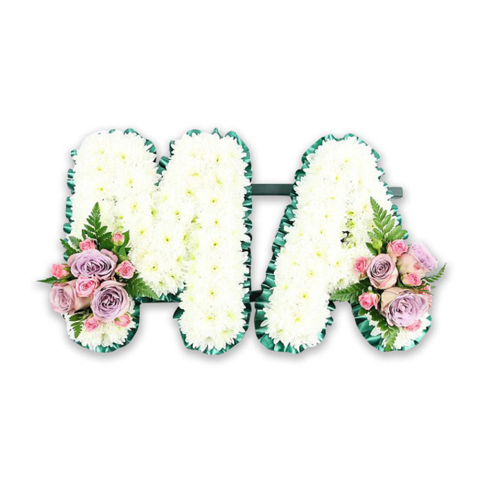 Photographed against a plain white backdrop, this floral tribute spells out the letters MA in bold, structured capitals, offering a heartfelt farewell likely intended for a beloved mum or mama. Each letter is generously covered in a tight mosaic of white chrysanthemums, the small, many-petalled blooms nestled closely together until they form a plush, almost quilted surface. No stems or gaps are visible, just a continuous field of soft white, its texture reminiscent of a cloud or a thick blanket of snow. This pure, uniform colouring brings a sense of calm, ideal for a funeral service in Collier Row where families seek a quiet, dignified expression of their affection. The edges of the letters are sharply defined by a deep green satin ribbon, neatly pleated and carefully pinned to follow every corner and curve. This rich emerald border provides striking contrast and frames the floral letters with a polished, finished look, ensuring that the M and A stand out clearly from any distance in the chapel or crematorium. At the lower left base of the M and the lower right base of the A, the design is enhanced with small, coordinated floral clusters. Here, soft-pink roses and delicate lilac roses are arranged together, some flowers more open, their spiral centres clearly visible, while others remain in bud, suggesting tenderness and unfolding love. Smaller pink spray roses dot the arrangement, filling in spaces with their petite blooms. A few light fern fronds arc outwards, joined by snippets of lush foliage, adding shape and a hint of garden freshness. The result is a balanced and deeply personal tribute, the kind of bespoke letter arrangement a local Collier Row florist might create as a final message of gratitude, love and remembrance.
