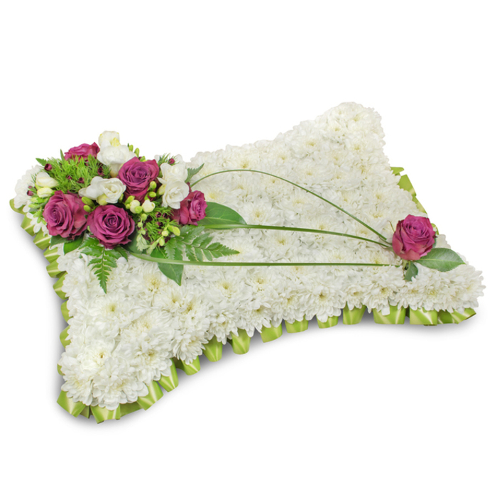 A carefully formed pillow tribute set against pristine white, lovingly constructed by Collier Row Flowers with a dense bed of white chrysanthemum florets that creates a plush, cloudlike texture meant to evoke peace and restful sleep. The perimeter is bordered by a pale lime-green satin ribbon, folded into a neat scalloped trim that frames the cushion and adds a soft, architectural finish. Dominating the upper-left quadrant is a richly coloured accent cluster of deep fuchsia-magent a roses, their velvety petals unfurling with gentle volume, punctuated by slender stems of white freesia or creamy lisianthus which add a fragrant, ethereal touch. Lush emerald foliage, including feathery fern fronds and smaller glossy leaves, provides contrast and a natural, living base for the blooms. Three slender bear-grass stems arc gracefully from the floral cluster across the pillow, their bright linear shapes introducing movement and guiding the eye toward the opposing corner where a single matching magenta rose is purposely placed among a few green leaves for compositional symmetry. The arrangement reads both solemn and tender: colour is restrained to bright greens, pure whites, and vivid magenta to convey respect and fond remembrance. You can imagine this tribute arriving at a Collier Row chapel, a family home near Collier Row Lane, or being laid at a service in Romford-fresh-scented, meticulously assembled, and intended to cradle memories with quiet dignity.