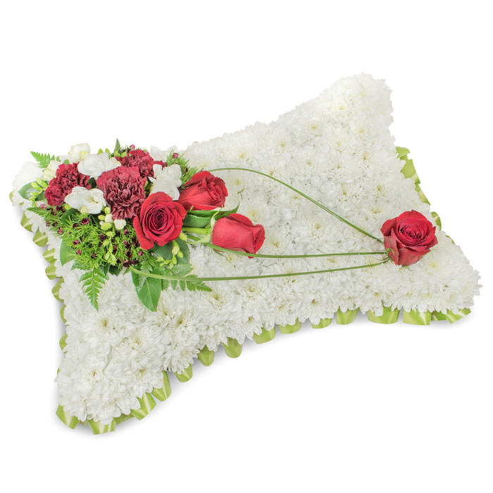 An expertly assembled funeral pillow shaped like a small rectangle, photographed against a clear white background so every detail is visible: the full surface is densely carpeted with immaculate white chrysanthemum heads, each bloom layered tightly to create a plush, cloud-like texture that suggests peace and rest. Along the slightly raised edge a pale green ribbon and slender foliage peep out, giving a neat, finished rim that softens the silhouette. On the upper left, an artful spray of deep, true-red roses and richly toned burgundy carnations forms the visual heart of the tribute; delicate white freesias and fine spray chrysanthemum buds are woven through this cluster, their lighter tones catching the eye and breaking up the strong reds. Behind these blooms, an underlay of rich green leaves and feathery fern fronds provides depth and contrast. From that corner a trio of elegant, curving bear-grass stems or similar bright green foliage extends diagonally across the white surface toward the lower right, creating a sweeping line that connects the main cluster to three scattered red roses, the final rose placed deliberately at the pillow's lower right-symbolic of enduring love carried across the arrangement. The scent is a muted wash of rose and fresh greenery, the lighting soft and contemplative, and the overall mood respectful and calming. This classic cushion style is often chosen by families in Collier Row Village, Romford and surrounding Havering districts when a traditional, timeless floral tribute is wanted for a service or memorial.
