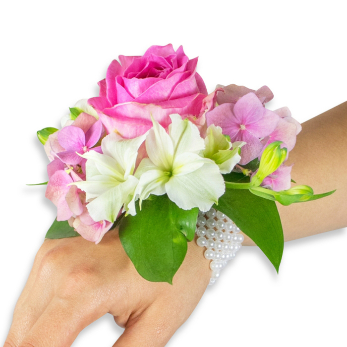 A refined, wearable bouquet shown on a left wrist, photographed on a clean white field to emphasize colour and form. The focal point is a large, fully bloomed deep fuchsia-pink rose whose concentric, velvety petals create a radiant center; surrounding it, soft blush-pink hydrangea florets build up a voluptuous, cloud-like texture that cushions the rose. Interspersed with the hydrangea are crisp white alstroemeria (Peruvian lilies), each presenting four to six smooth petals with delicate pale green streaks at their throats, lending botanical detail and lightness. To the right of the rose a tasteful cluster of pale green alstroemeria buds and a few small, barely opened pink blooms hint at further life and motion within the arrangement. Broad, glossy emerald leaves cradle and contrast the bouquet, defining edges and adding depth against the wearer's light-to-medium skin tone. The floral cluster is affixed to the wrist by an elegant, multi-strand bracelet of small white pearls that glimmer subtly as they wrap around the wrist and trail partway onto the back of the hand, combining comfort with old-world charm. As a local touch, this piece reflects the understated elegance preferred by Collier Row residents for weddings, proms and community galas near Romford - and at Collier Row Flowers we prepare corsages like this to be fresh, wearable and thoughtfully balanced, available for local delivery across Collier Row, Romford and surrounding streets when needed for special occasions.