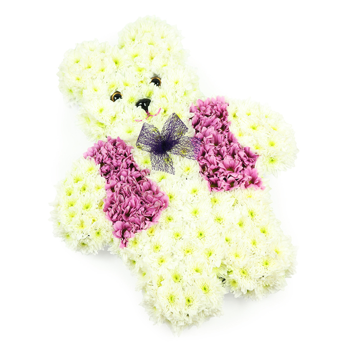 Shown against a pristine white backdrop, this floral teddy bear tribute appears both tender and carefully composed, as if it has been shaped by hands that understand how flowers can speak when words are difficult. The bear itself is constructed from numerous small chrysanthemums in shades of creamy white and very pale yellow, each flower head set closely against the next to create a dense, plush surface. The contours of the head, rounded ears, and softly padded paws are defined by the way the blooms follow the curves, enhanced by subtle shadows from the gentle, even lighting. Small, dark button-style eyes and a neat triangular nose are positioned with care, giving the bear a soft, reassuring gaze that feels especially touching for a child's remembrance. Draped across the chest is a floral waistcoat made of rich pink and magenta chrysanthemums, the deeper tones standing out vividly from the lighter body, much like the bright floral touches seen along Collier Row's residential streets in late spring. The waistcoat is shaped into neat, lapel-like sections that meet at the centre of the chest. Just at that point rests a delicate bow crafted from sheer purple netting, its translucent loops adding fine, gossamer texture and a whisper of playfulness. No foliage or extra decoration distracts from the clarity of the design; the bear stands alone, a clear symbol of comfort and love. As a funeral or memorial tribute for families in Collier Row, this floral teddy offers a poignant blend of innocence, beauty, and quiet solace.